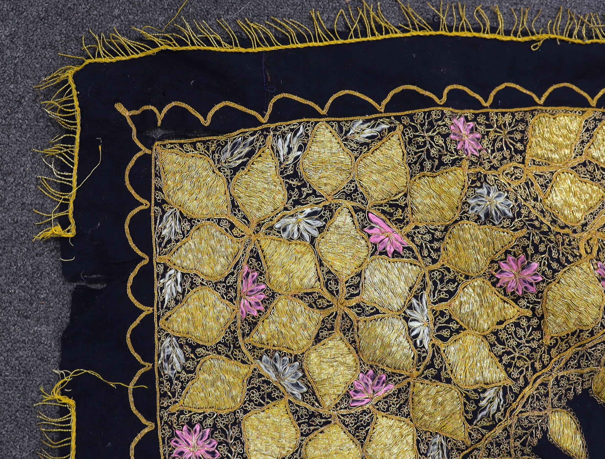 An Indian gold thread and coloured silk floss embroidered black wool cover with gold thread fringing and a later woven cover with bobble edging, black and gold cover 172cm x 169cm
